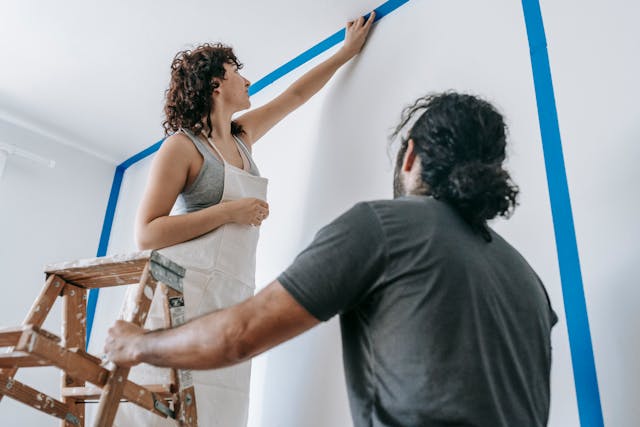 Person standing on a ladder painting a wall while another person holds the ladder